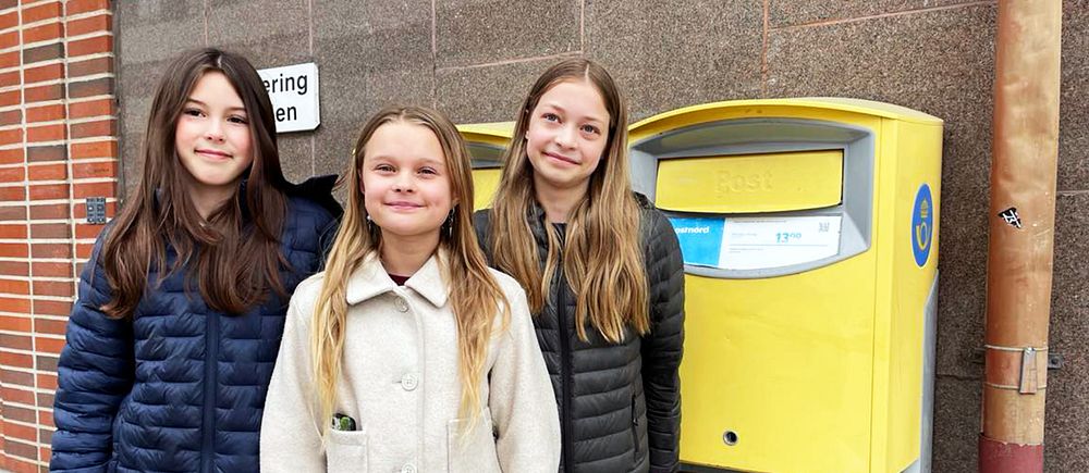 Tre personer står framför en gul brevlåda vid en tegelvägg.