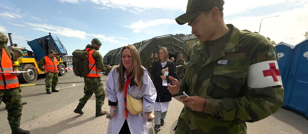 Militär personal och sjukvårdare samarbetar utomhus vid ett akutområde med tält och fordon.