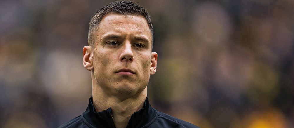 AIK-spelare med mörkt jacka och klubbmärke synligt, suddig publik i bakgrunden.