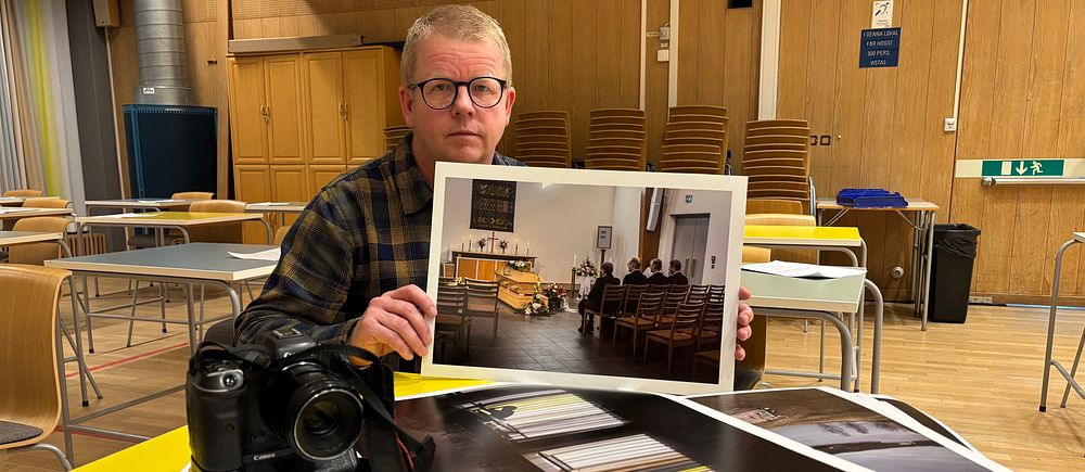 Person visar fotoutskrifter vid bord med kamera. Bord och stolar syns i bakgrunden.