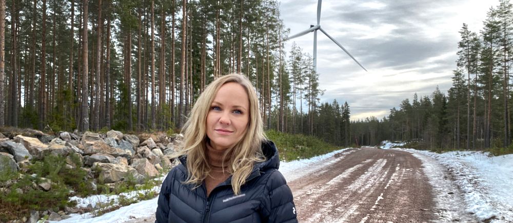 Kvinna i mörk jacka på en grusväg vid vindkraftverk och tallskog.