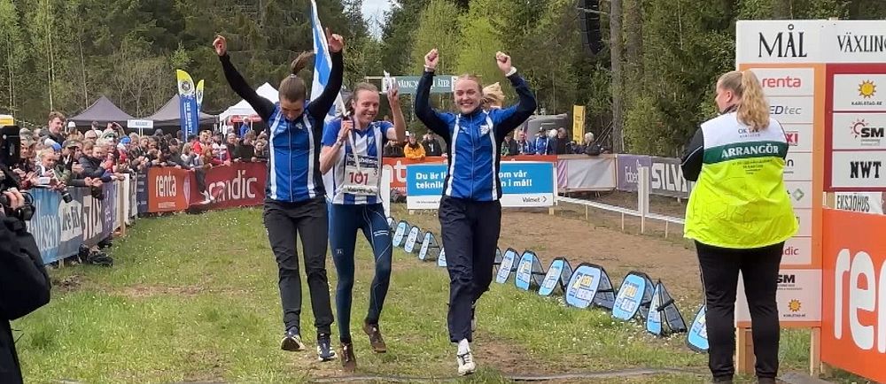 Tre idrottare firar med armarna i luften vid en mållinje, publik hejar längs sidorna.