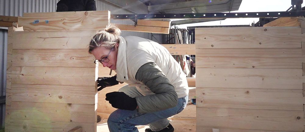 Kvinna i vinterkläder böjer sig ned omgiven av trälämmar i ett byggnadsområde där en förening i Göteborg bygger en rullande bastu som ska vara gratis för alla att använda.