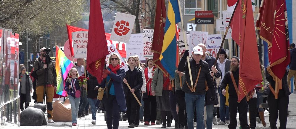 Demonstrationståg med svenska flaggan och röda fanor, människor bär plakat och skyltar.