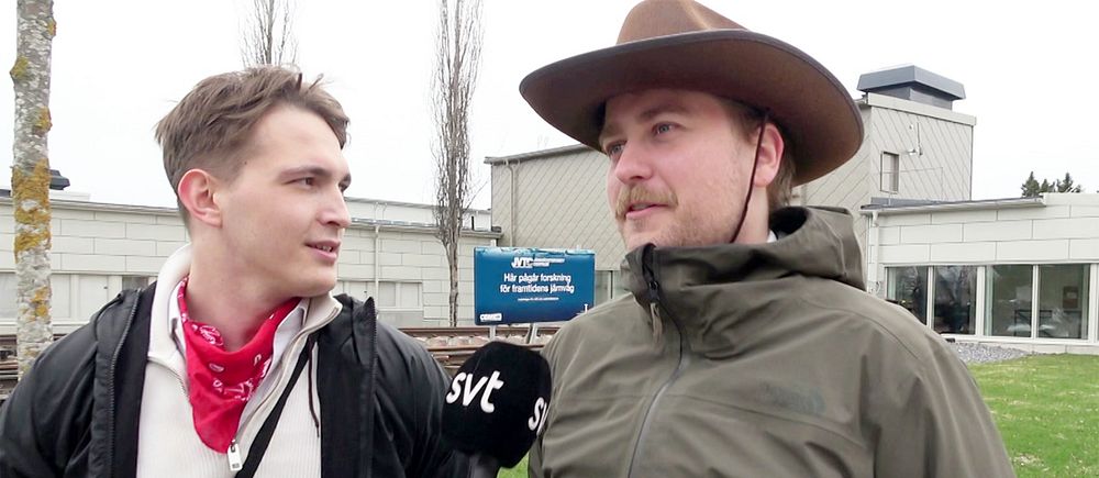Två personer intervjuas utanför en byggnad. En av dem har en cowboyhatt på huvudet.