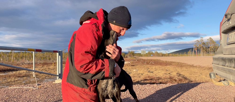 Man i röd jacka kramar en hund på grusväg under blå himmel.