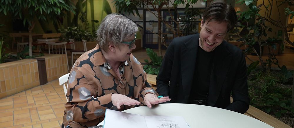 Två personer vid ett bord, diskuterar en bok med illustrationer.