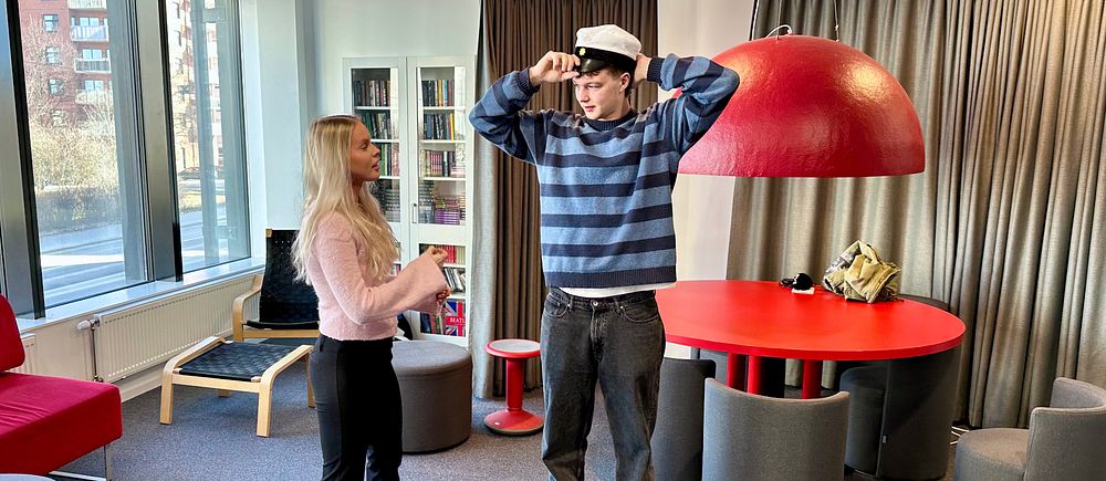 Två personer står i ett färgglatt rum, en man tar av sig en studentmössa.