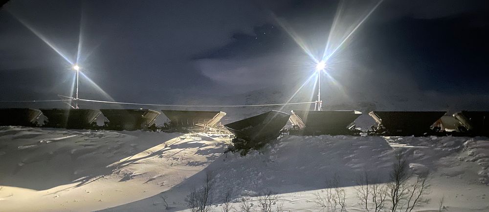 strålkastare över malmvagnar på vall, snötäckt mark. En vagn hänger ner längs vallen