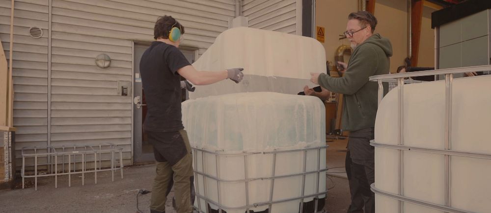 Två personer hanterar en stor  plastbehållare med is i en industrilokal.