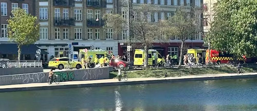 Flera ambulanser står parkerade vid kanalen framför en lägenhetsbyggnad i stadsmiljö.