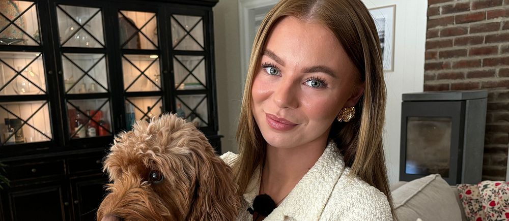 Kvinna med tatuerad arm sitter i ett vardagsrum och håller en brun hund i knät.