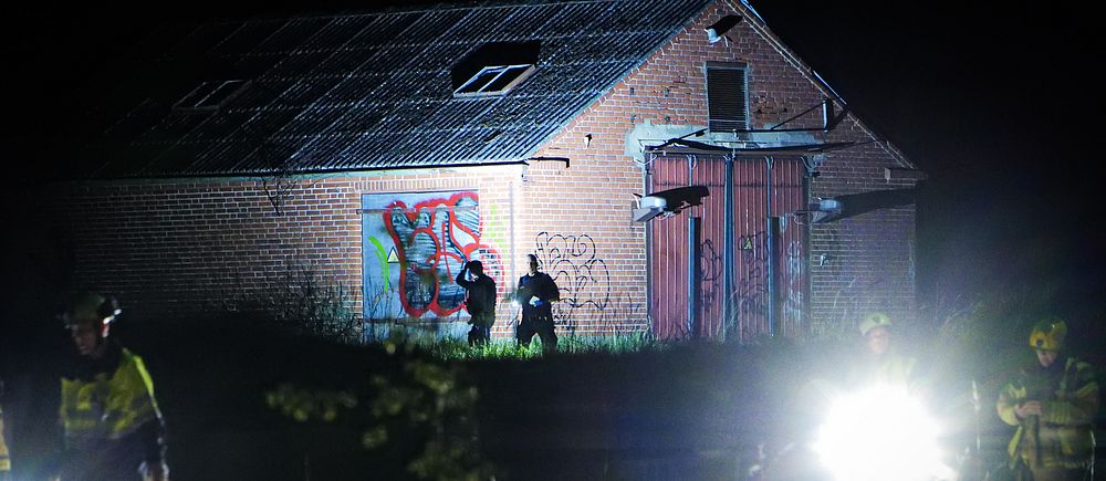 Polis undersöker området vid en nedgången lada nattetid, stark belysning i förgrunden.