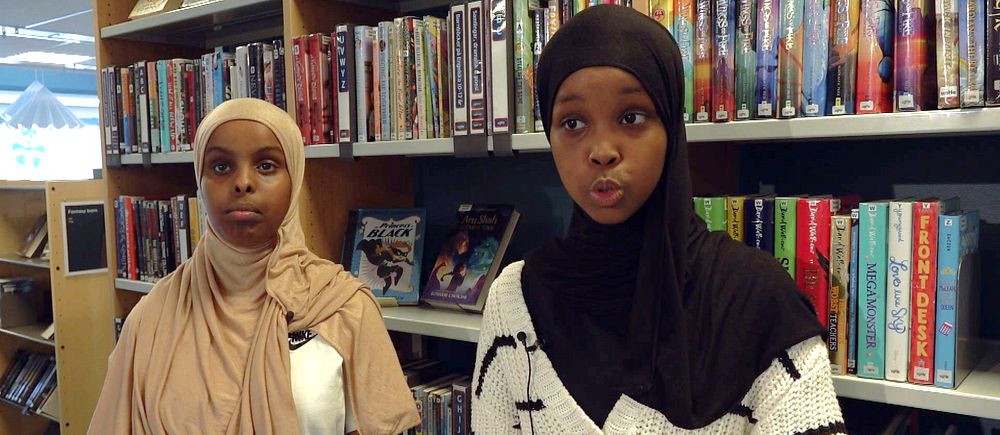Två tjejer med hijab framför hylla med böcker i bibliotek