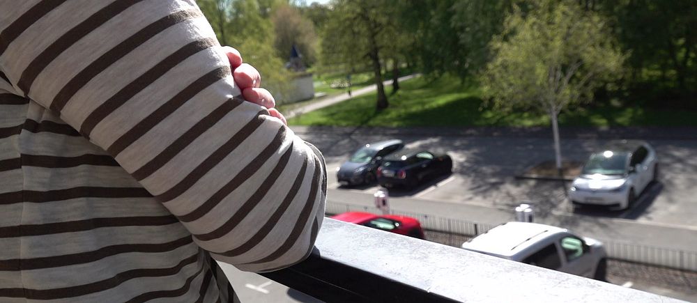 Person i randig tröja lutar sig mot balkongräcke och tittar ner mot parkering och grönområde.