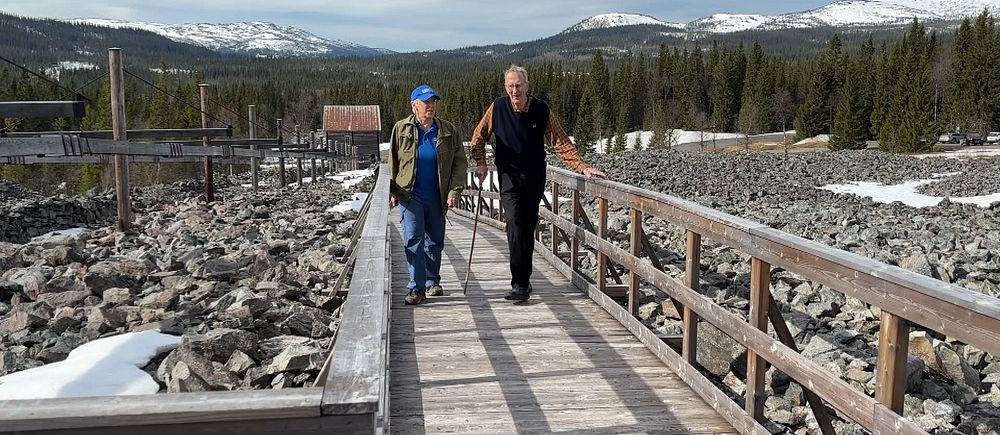 Två personer promenerar på en träbro i Fröå gruva med snö och berg i bakgrunden.