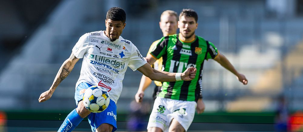 Fotbollsspelare i vit och blå dräkt sparkar bollen, motståndare i grönsvart förföljer.