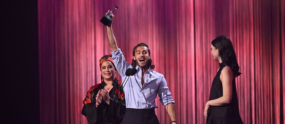Man firar med pris, omgiven av två kvinnor på scen under en gala.
