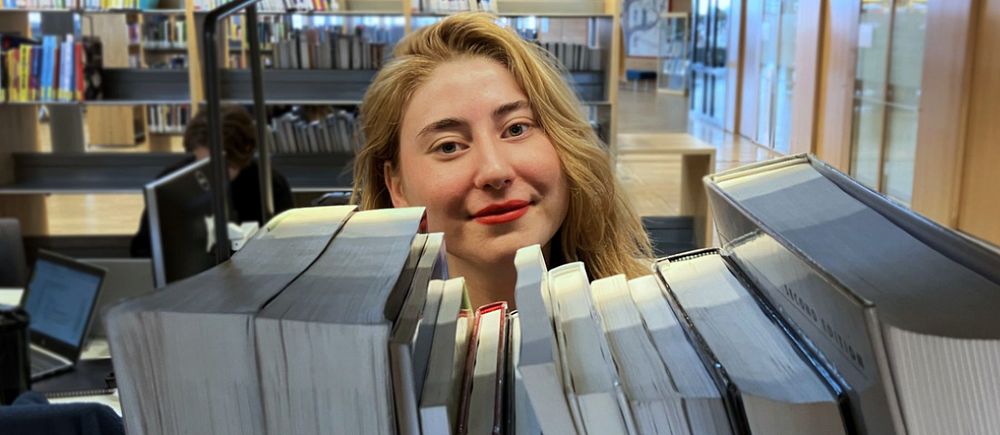 Kvinna med ljust hår bakom böcker i bibliotek, Olivia Rosén vid Karlstads universitet, vinnare av Lilla Psykologipriset 2024