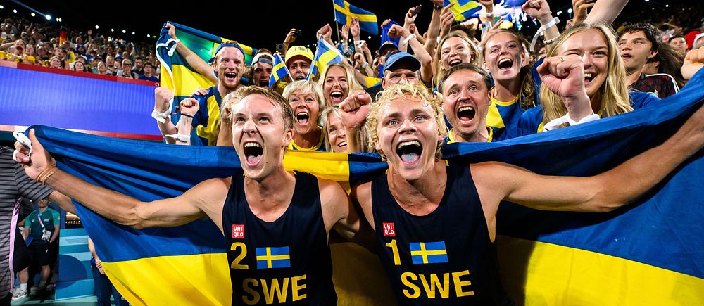 Glad grupp svenska idrottare och fans som firar med flaggor på stadion. Jonatan Hellvig och David Åhman.