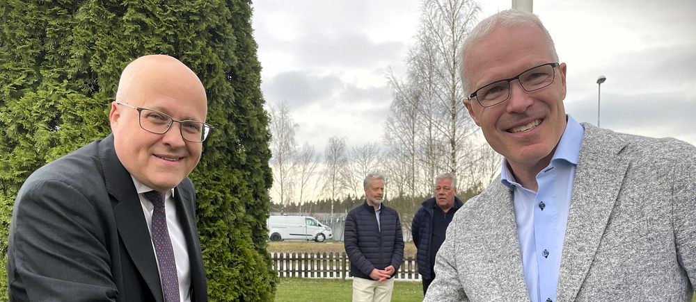 Två personer skakar hand utomhus, observation av två åskådare i bakgrunden.