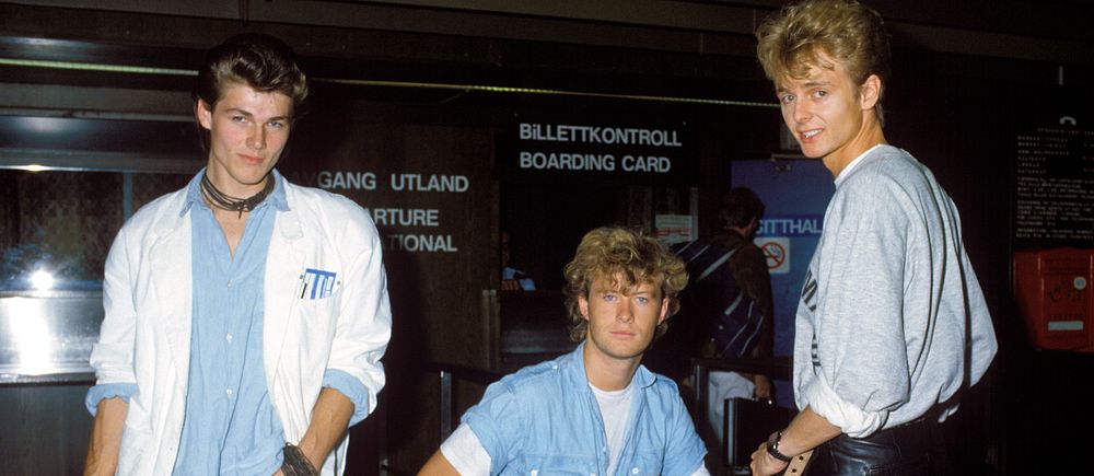 Tre unga vuxna i 1980-talskläder står vid en biljettkontroll på en flygplats.