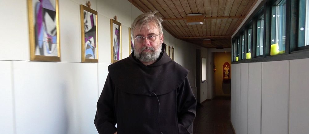 Person i mörk dräkt går i korridor med tavlor på väggen och ljusinsläpp från fönster.