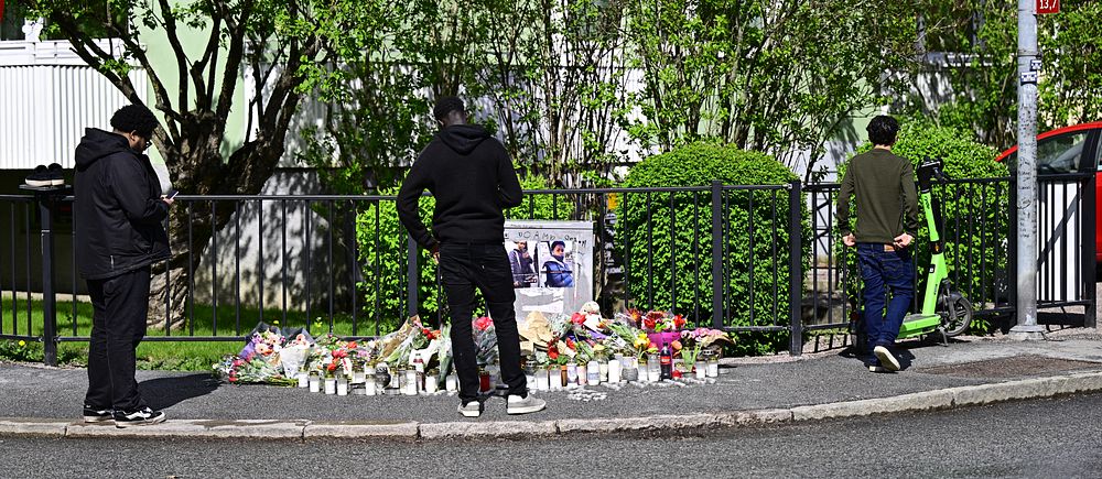 Personer samlas vid en minnesplats med blommor och ljus vid trottoarkant.