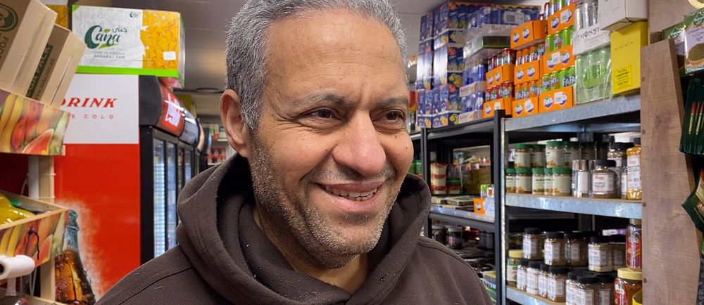 Ameen Talal står i sin butik i Lagersberg, som . Bakom honom syns hyllor med konserver och läskburkar. Han ser glad ut.