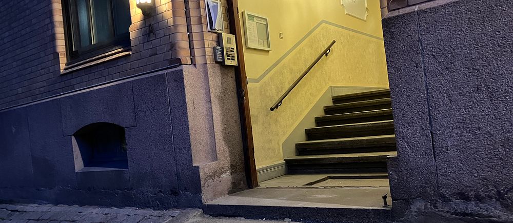 Entré till en byggnad med upplyst trapphuset på kvällstid. Trappor leder in till dörren.