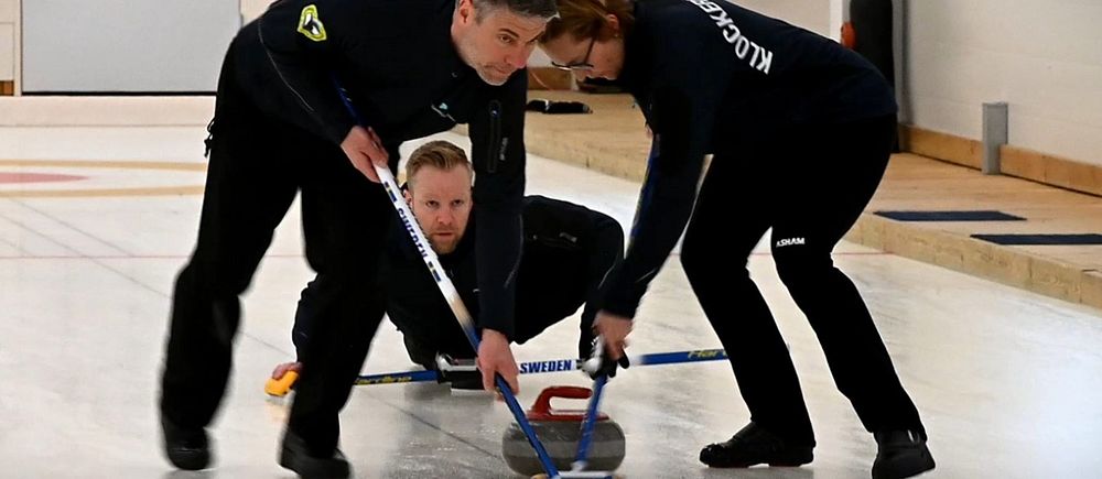 Tre personer spelar curling, två sopar framför stenen medan en släpper iväg den på isen.