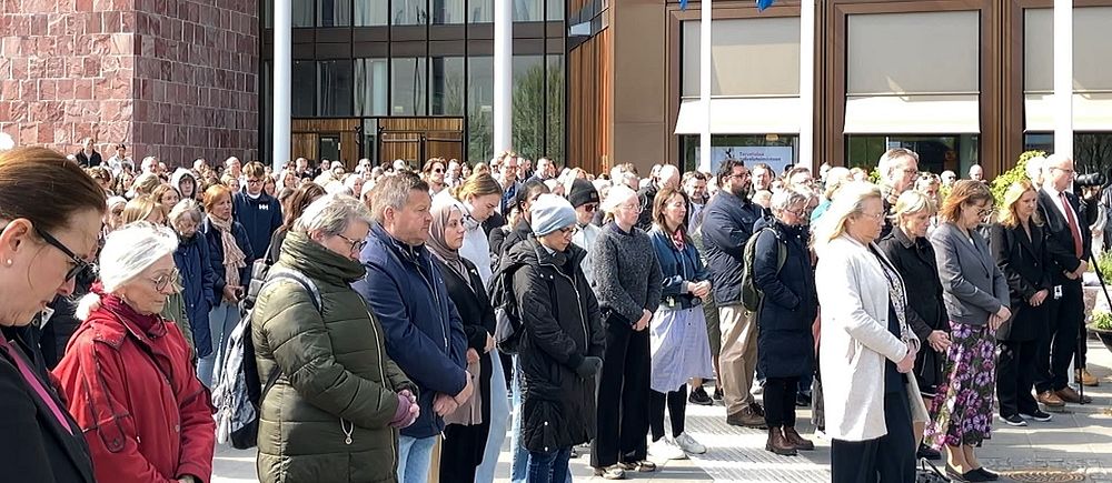 Stor folkmassa står samlad utanför en byggnad med svenska flaggor i solsken.