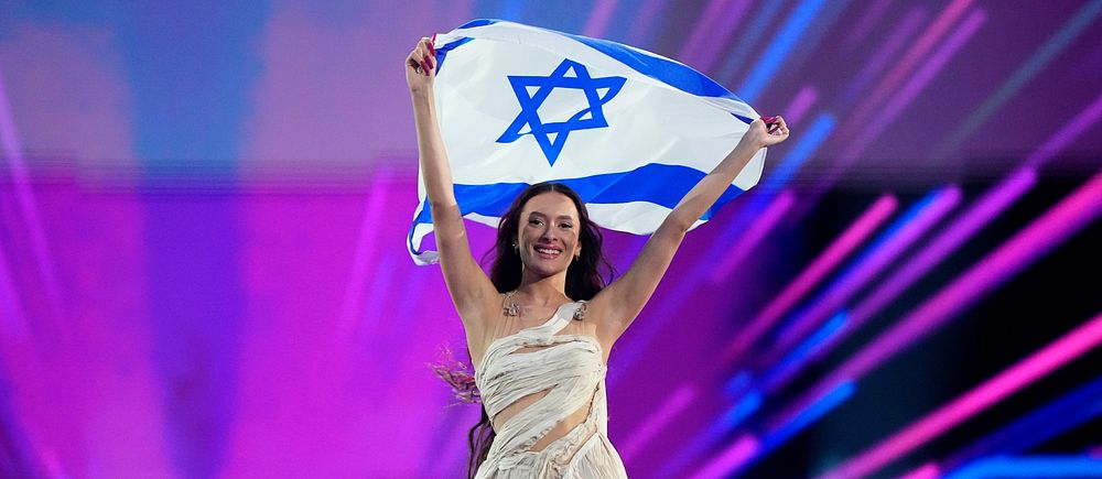 Kvinna i vit klänning håller Israels flagga på scen med färgglada ljuseffekter.