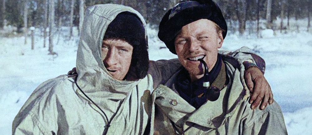Två personer i vinterkläder står tillsammans i ett snöigt skogslandskap.