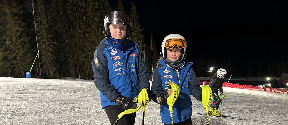 Två unga skidåkare från Almåsa alpina i står i skidbacken på kvällen.