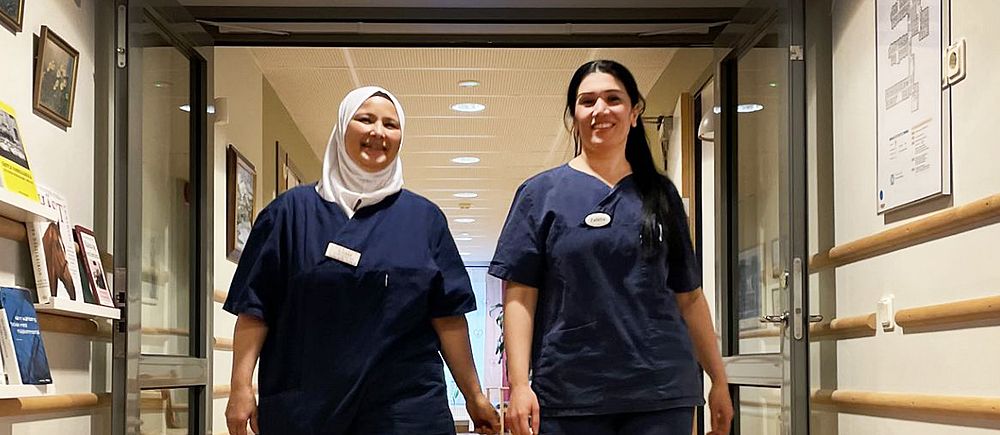 Kvinnorna Ilham och Zalikha i blå arbetskläder går i en korridor på ett äldreboende i Säffle