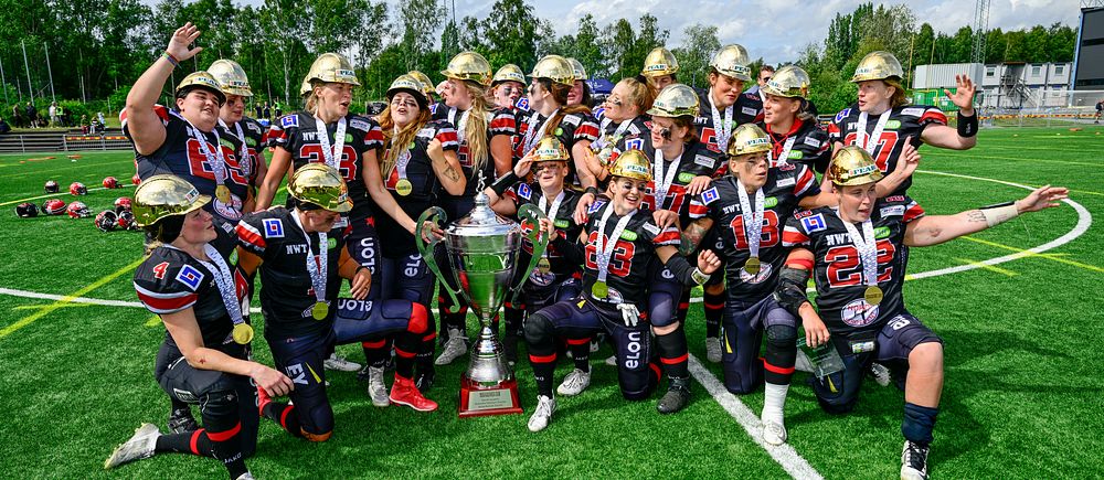 Carlstad Crusaders damlag iföda amerikansk fotbollsutrustning poserar med stora pokaler på en fotbollsplan.