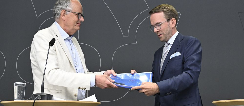 Två män i kostym överlämnar ett dokument vid ett podium under en formell presentation.
