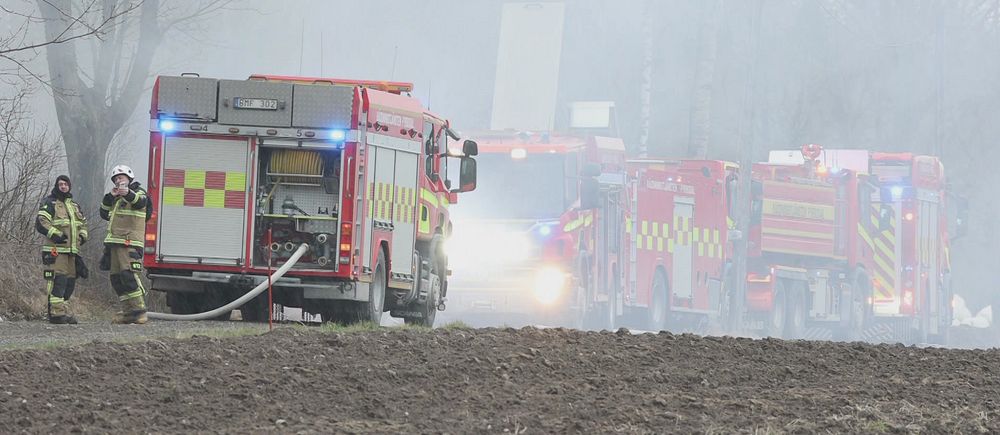 Brandbilar och brandmän vid rökfyllt område på landsväg nära åker.