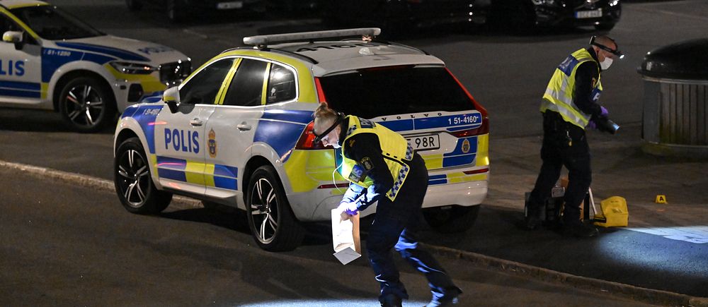 Poliser undersöker en plats med ficklampor vid en parkerad polisbil på kvällen.