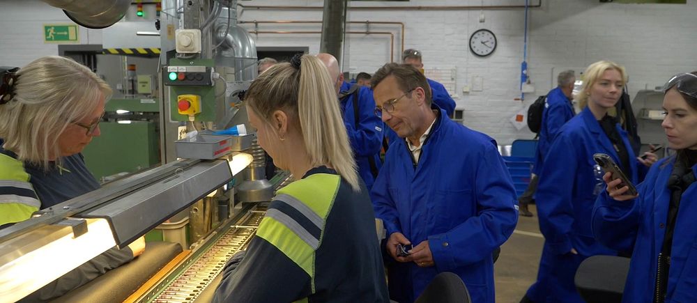 Personer i blå arbetskläder besöker en industrihall och studerar en pågående produktion.