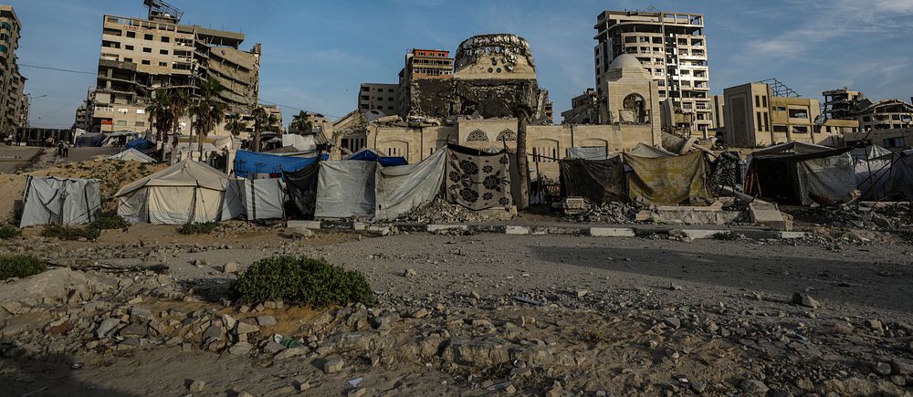 Sönderbombade byggnader och tillfälliga tält framför ruiner under blå himmel.