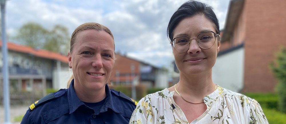 Två personer står utomhus, en i polisuniform och en i mönstrad blus. Hus syns i bakgrunden.