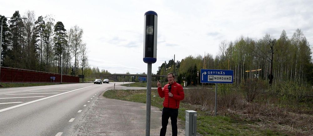 Reporter står med röd jacka och pekar på en fartkamera intill en väg.