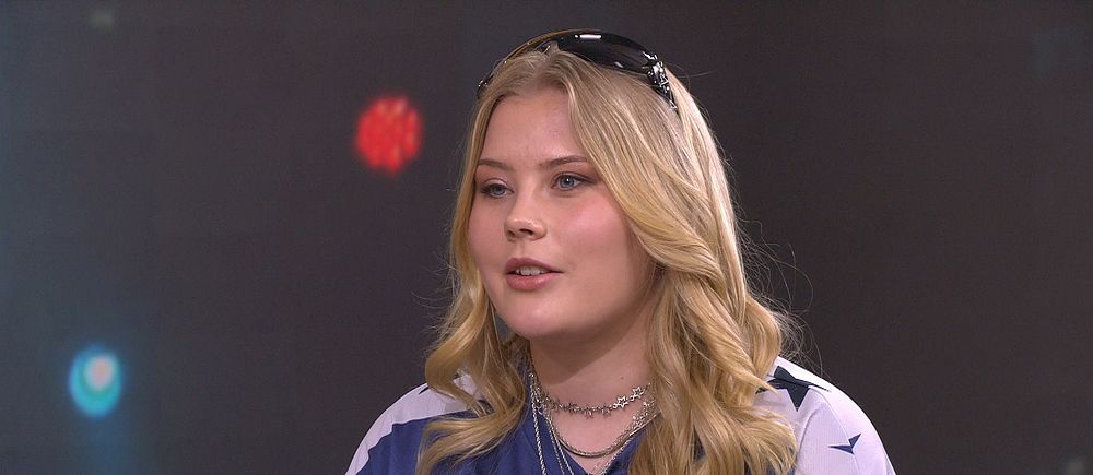 Person med långt blont hår och blå tröja med vita detaljer i studiomiljö.