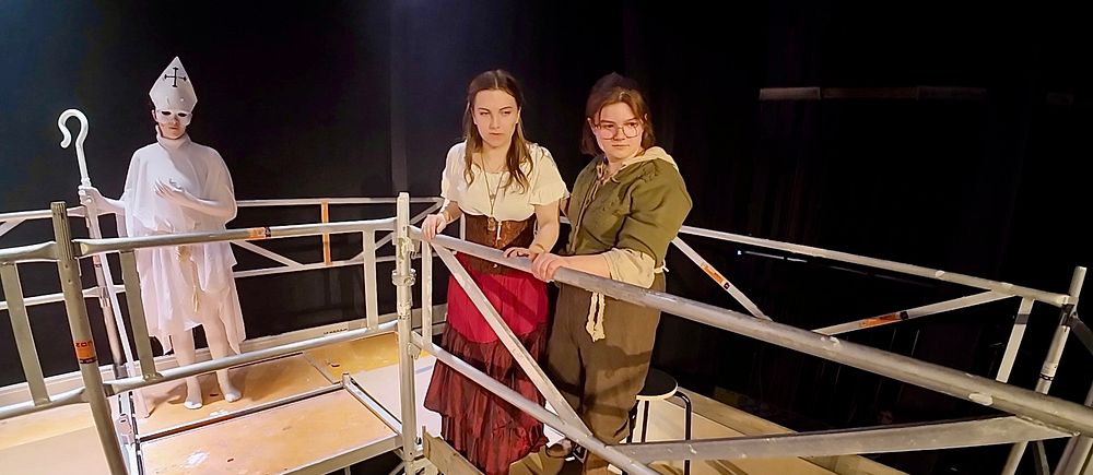 Två kvinnor i historiska kläder står på en teaterscen, en person i vit kostym syns i bakgrunden.