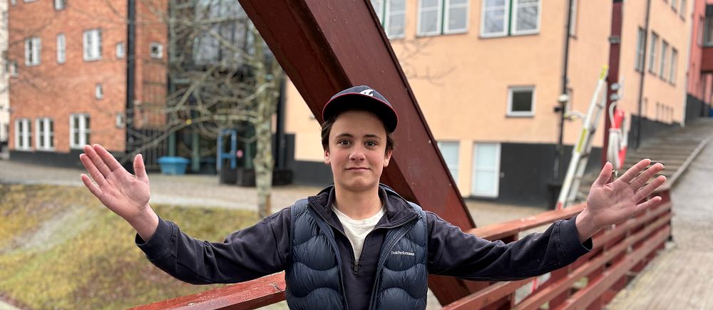 Person med keps står på bro med utsträckta armar, byggnader syns i bakgrunden.