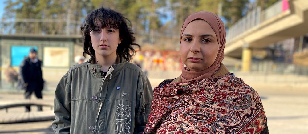 Två personer står ute bredvid en bro, bakgrund med träd och en busstation.