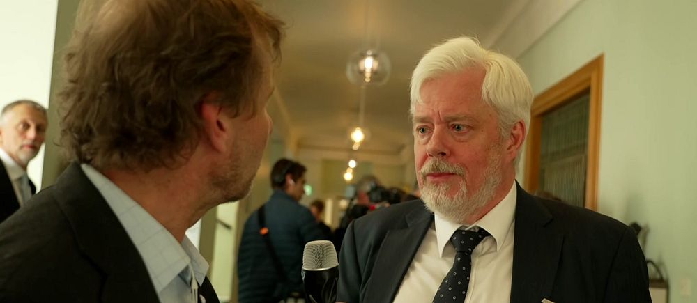 Reporter intervjuar Niklas Ekvall i kostym i en korridor, flera personer syns i bakgrunden.