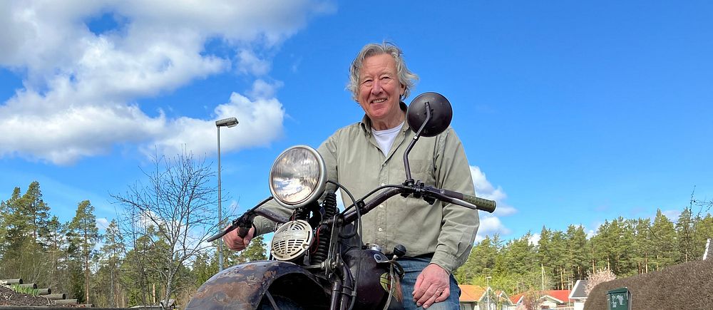 Leende man sitter på en motorcykel på en landsväg med skog och blå himmel i bakgrunden.